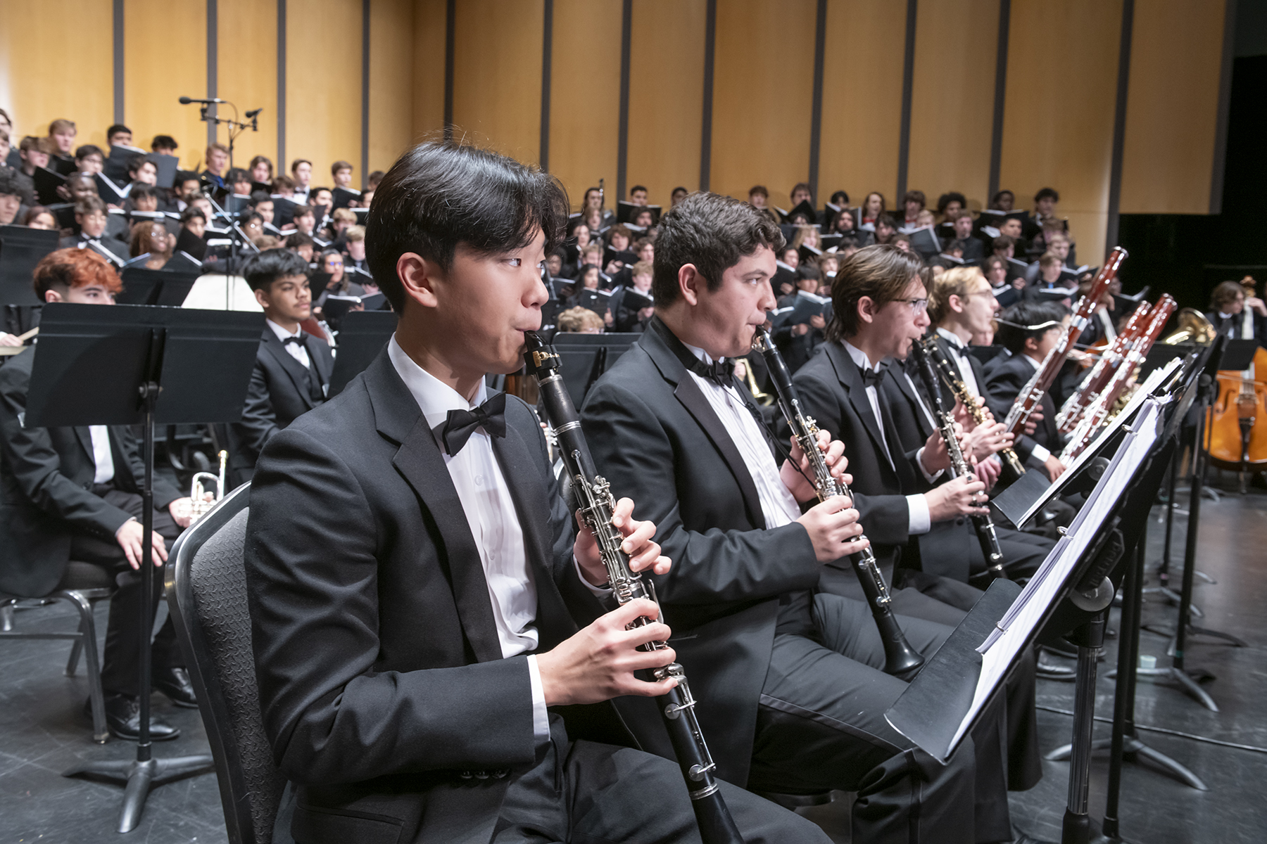 An instructor demonstrates violin technique to an attendee, assisting with finger placement on the instrument. The background is filled with a focused audience observing the demonstration, suggesting an educational workshop setting.