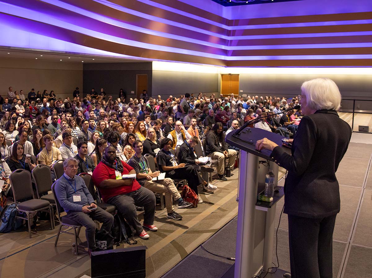 A speaker at a podium addresses a large audience in a conference hall with colorful lighting overhead. The crowd seems to be composed of professionals, gathered for a conference presentation.