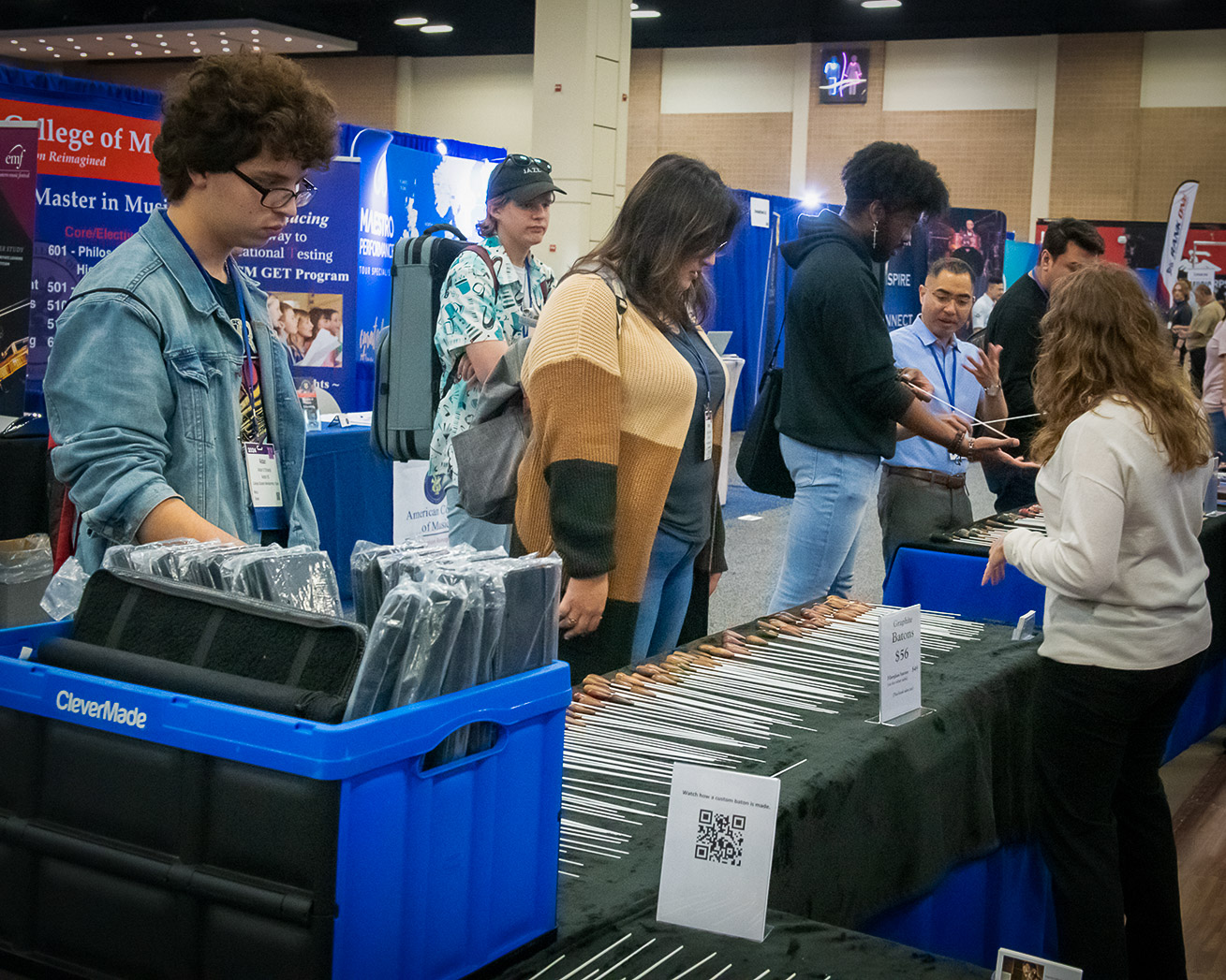 An instructor demonstrates violin technique to an attendee, assisting with finger placement on the instrument. The background is filled with a focused audience observing the demonstration, suggesting an educational workshop setting.